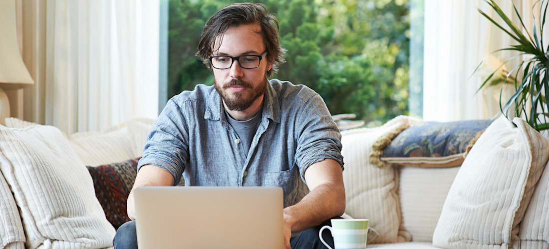 [Feature Image] A dark-haired learner wearing glasses researches the best way to learn Python programming on their laptop computer while sitting on a couch with a window displaying greenery behind them.