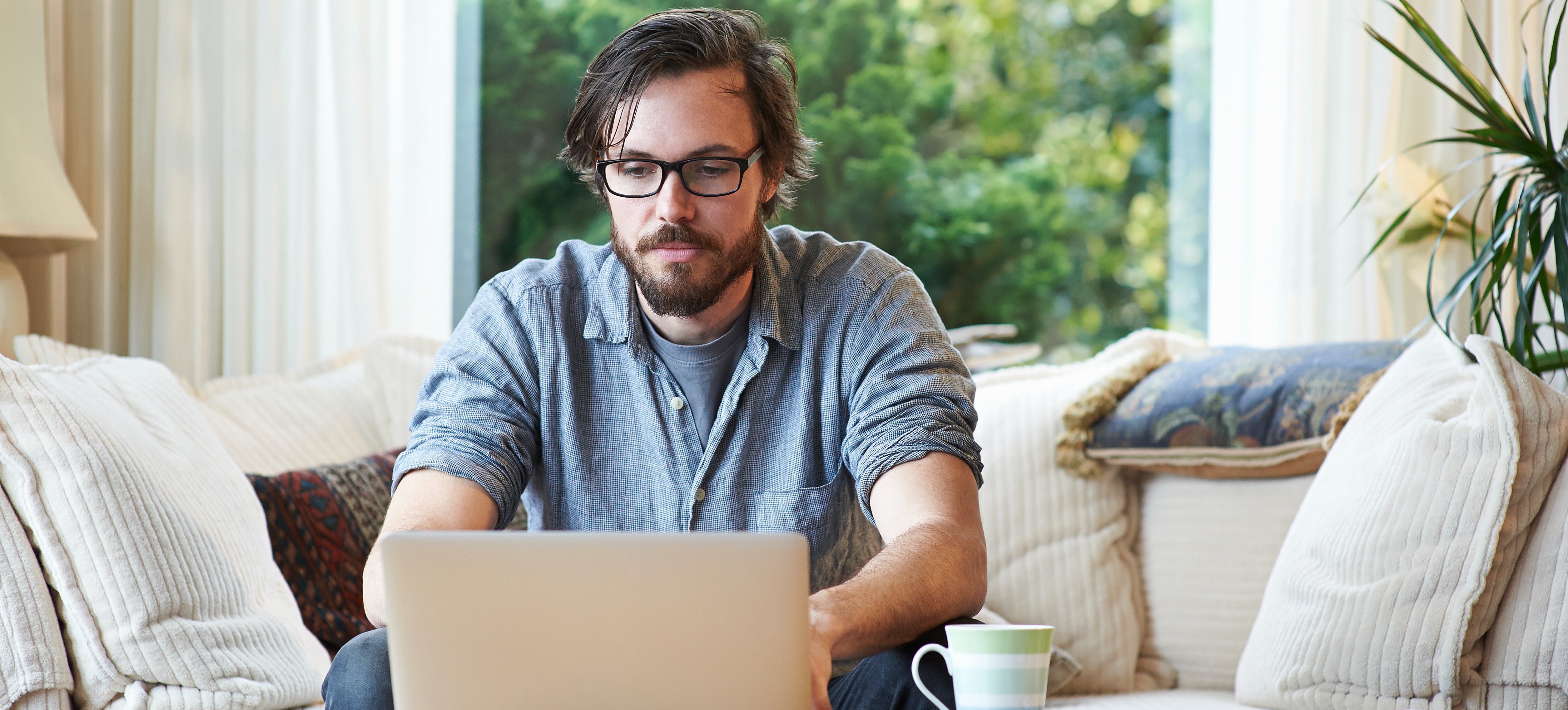 [Feature Image] A dark-haired learner wearing glasses researches the best way to learn Python programming on their laptop computer while sitting on a couch with a window displaying greenery behind them.