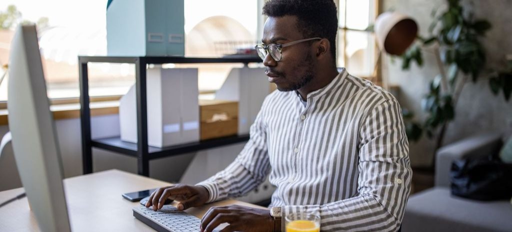 [Featured Image] An MBA student is wearing a striped button-up and glasses at their home office using a desktop computer. 