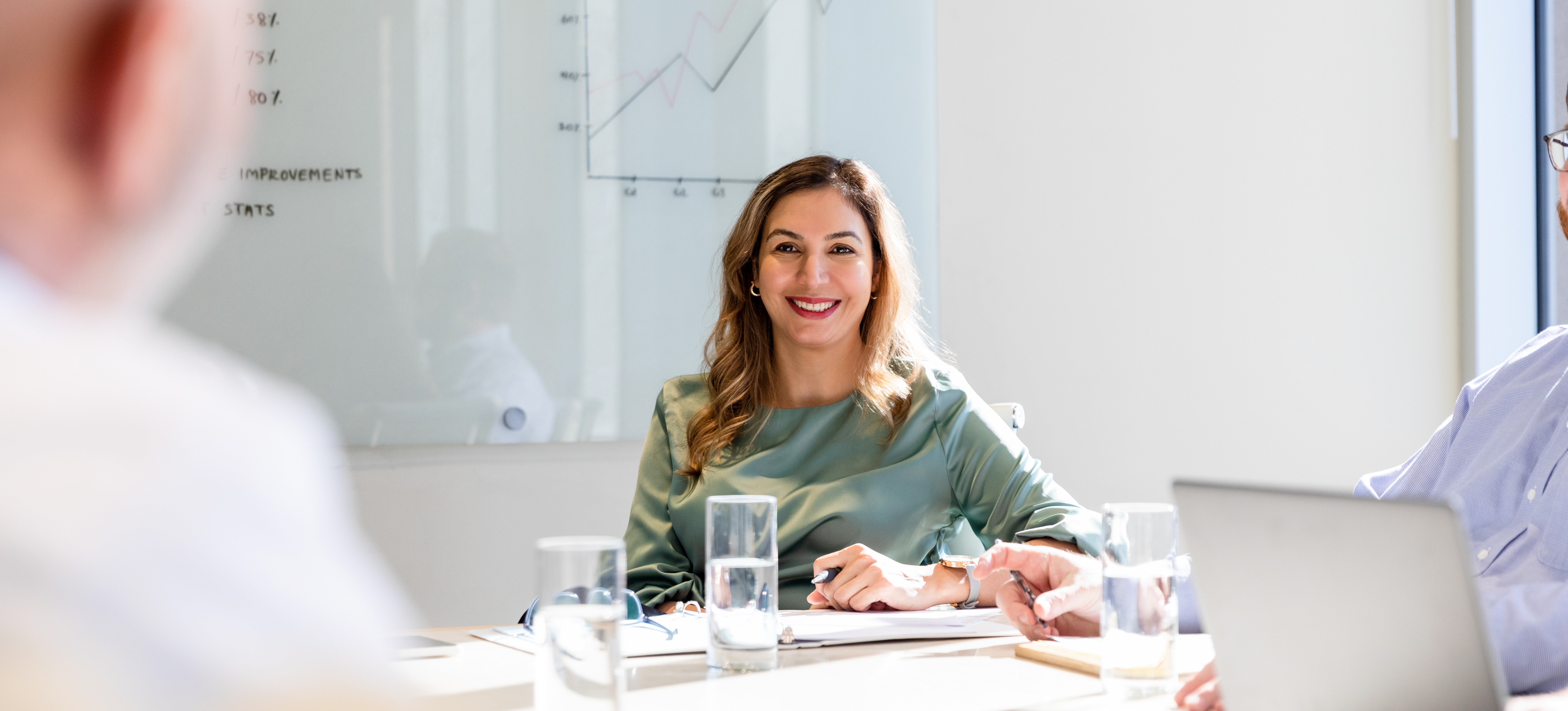 [Featured Image] A smiling woman CEO, one of the highest-paying business jobs, sits at the head of a boardroom table with a graph behind her, talking to two of her colleagues.
