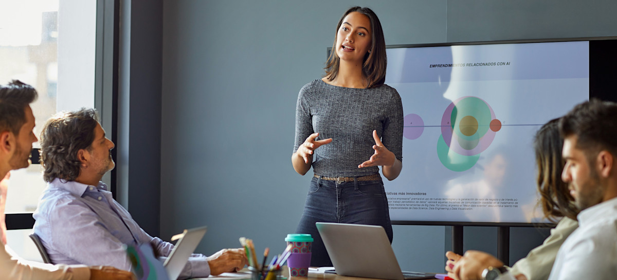 [Featured Image] A person working for an operations analyst salary stands before a group in a meeting space and presents in front of a screen filled with a graphic.

