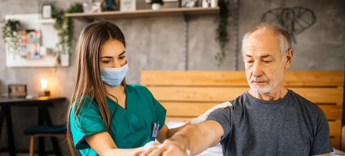 [Featured Image]:  A home health aide, with long brown hair,  wearing a green uniform and a blue face covering, is taking care of a patient in their home.  