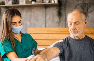 [Featured Image]:  A female home health aide, with long brown hair,  wearing a green uniform and a blue face covering, is taking care of a male patient in his home.  