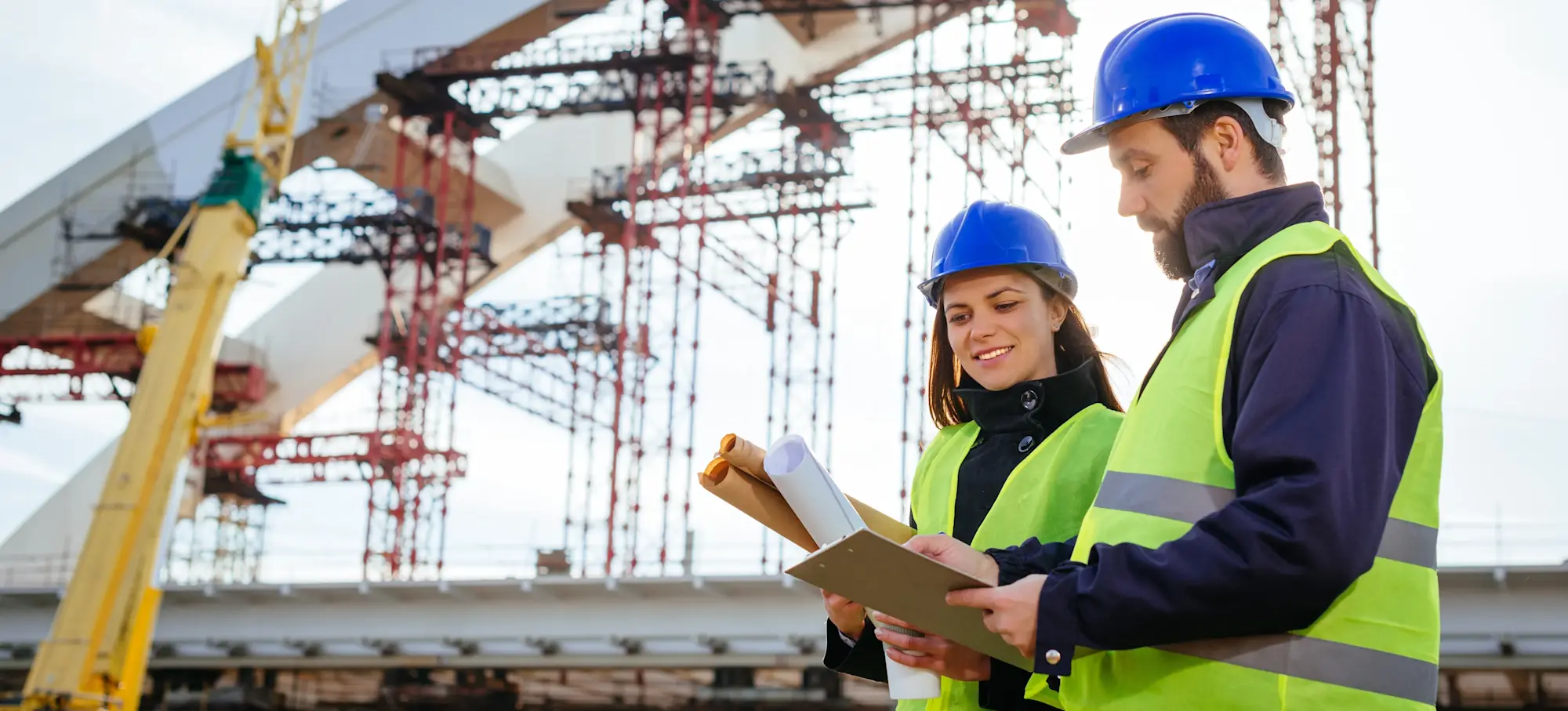 [Featured image] Two engineers in blue hard hats and neon vests work together on a construction site while looking at blueprints.