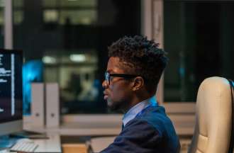 [Featured Image]:  A male wearing a blue sweater, blue shirt, and glasses, is sitting in front of his desktop, performing his duties as a data analyst. 