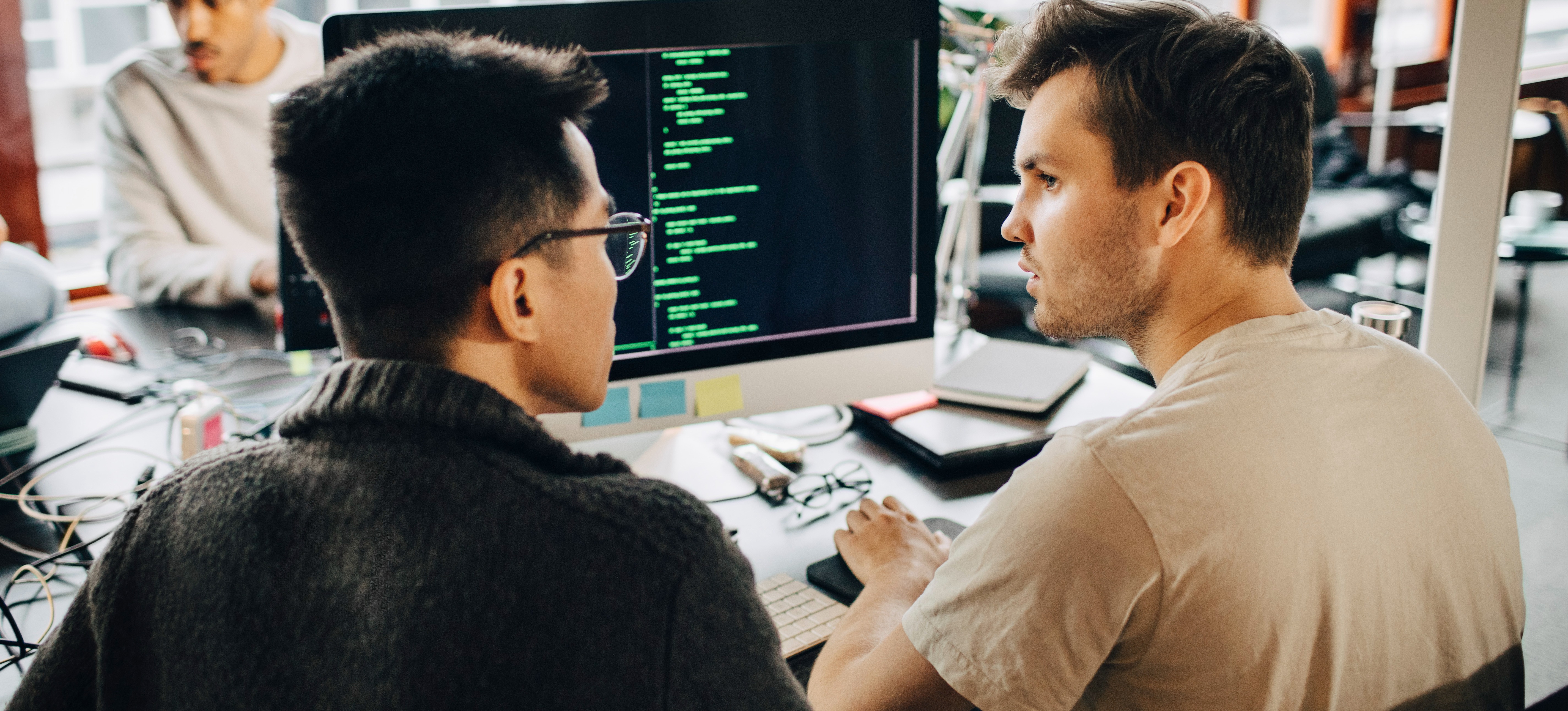 [Featured image] Two cybersecurity analysts sit in front of a computer monitor with code on the screen in a bright office.