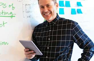 [Featured image] A man holding a tablet stands in front of a whiteboard where the 4 Ps of marketing are listed in green marker.