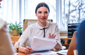 [Featured image] A white woman dressed professionally sits at her desk meeting with two people.