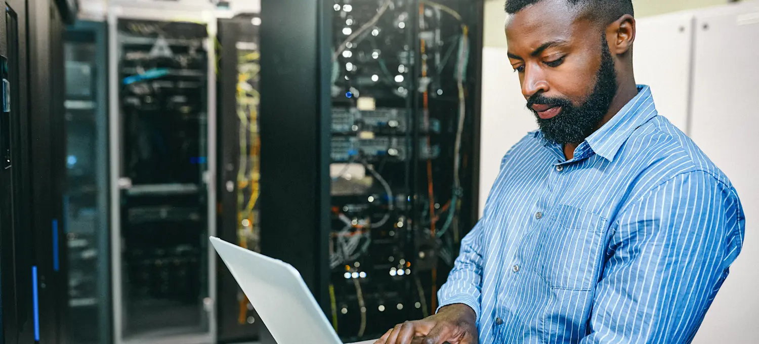 [Featured Image] A technical support engineer stands in a server room and looks at a laptop in an attempt to fix a problem. 
