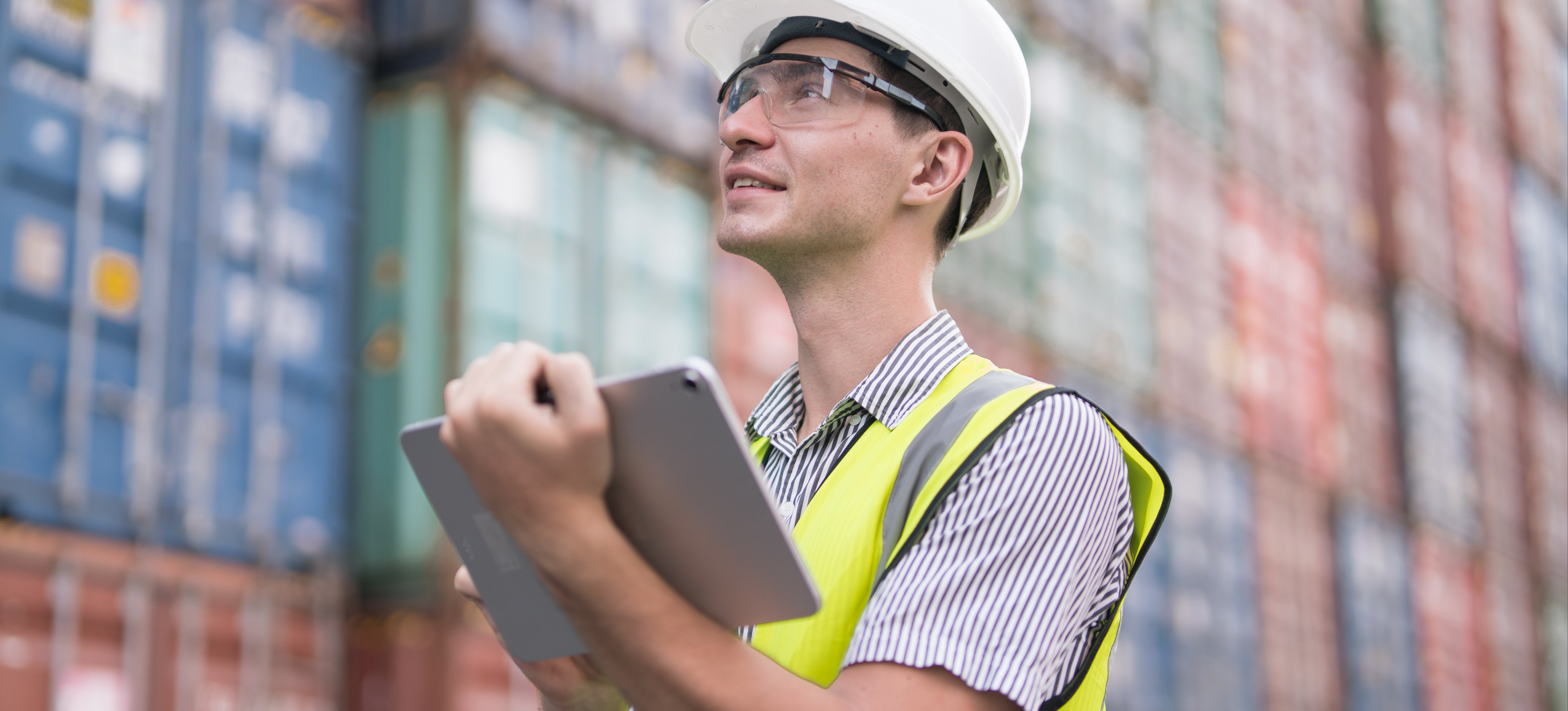 [Featured Image] A logistician wearing protective clothing and holding a tablet observes cargo freight in a container yard.

