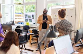 [Featured Image] A team of colleagues in a brightly lit room listening as one person speaks and uses data storytelling to bring data-driven insights to life.