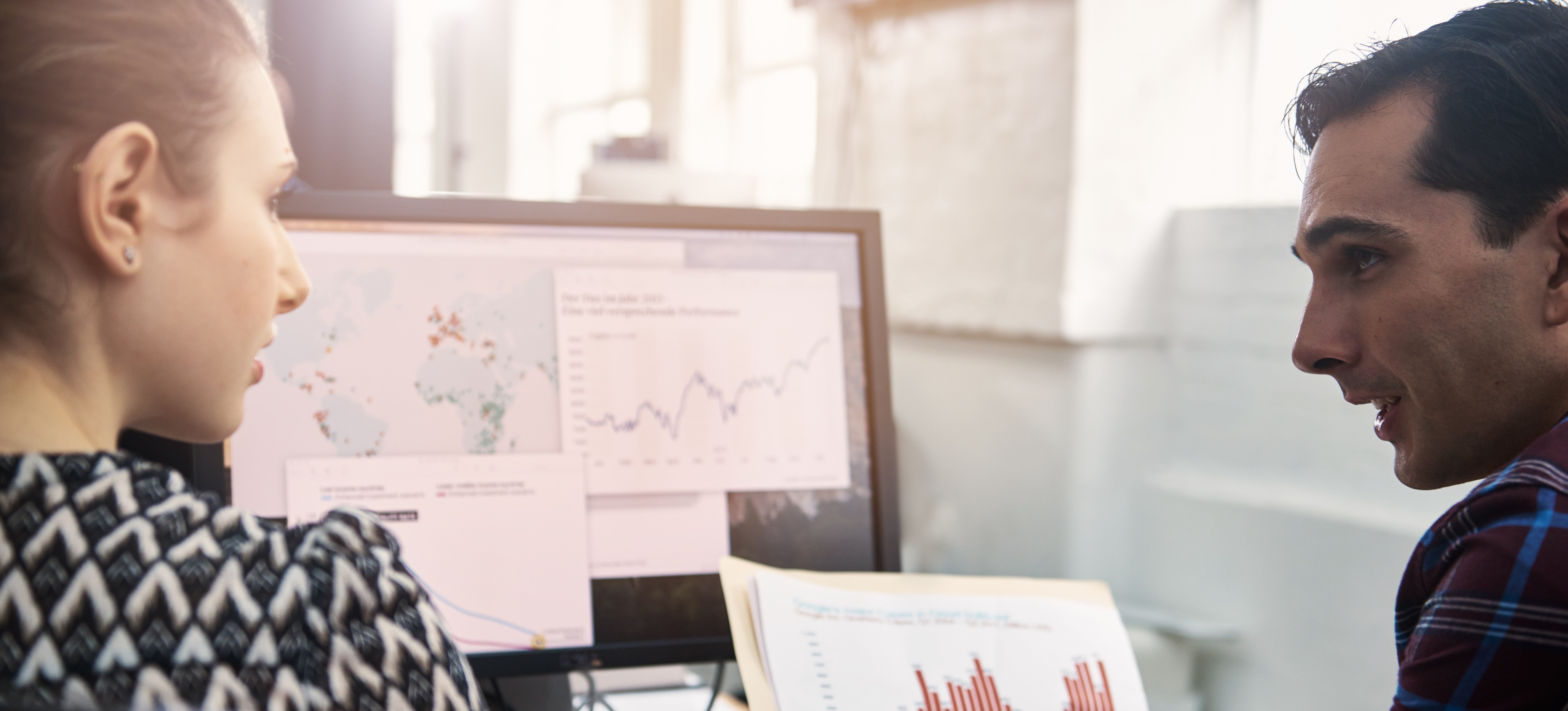 [Hauptbild] Zwei Datenanalysten diskutieren die Ergebnisse, während sie Datenvisualisierungen auf einem Computermonitor und Dokumente betrachten.
