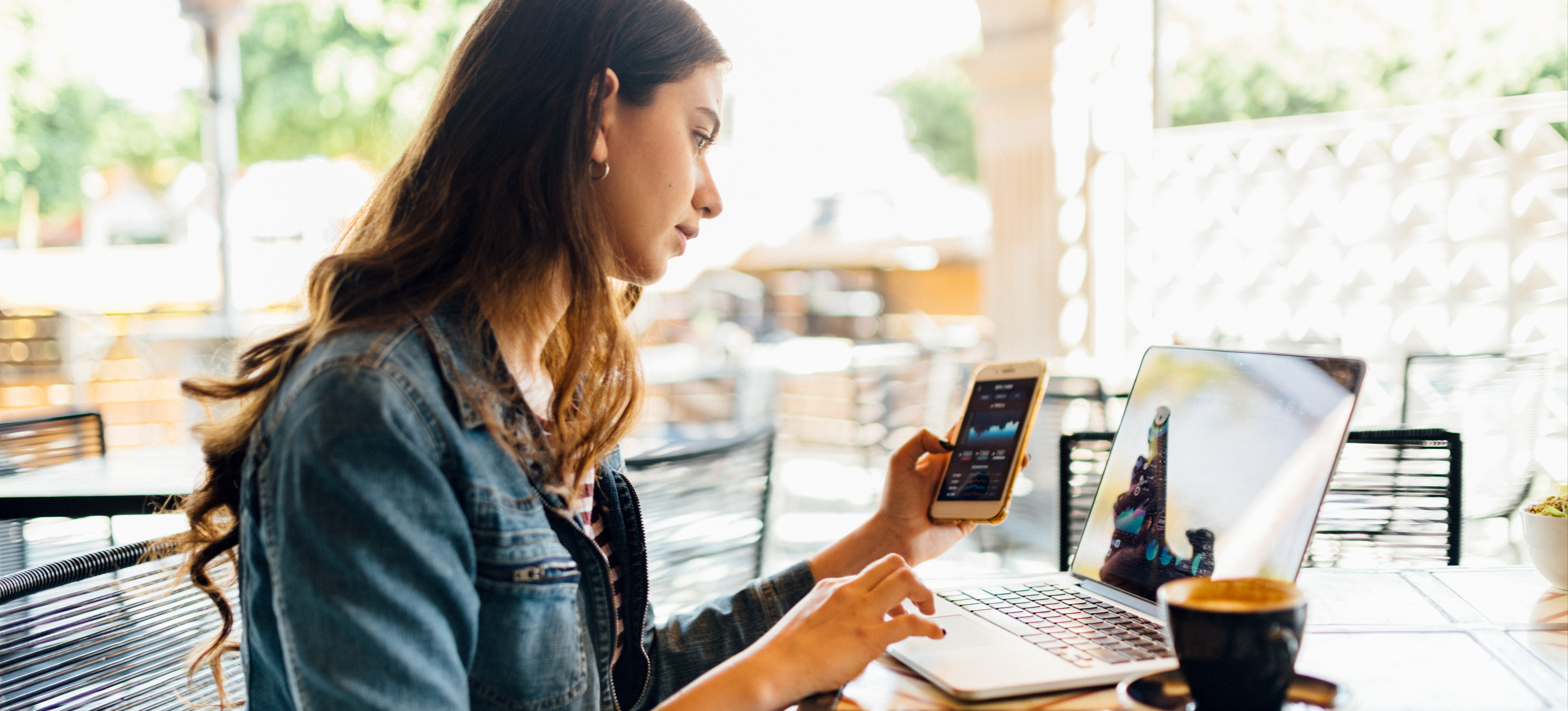 [Featured Image] A woman working on a computer and phone simultaneously in a coffee shop uses blockchain technology to monitor her crypto assets.
