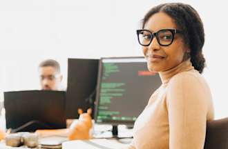 [Featured Image] A system administrator in a yellow turtleneck and black glasses sits at a shared desk with computer code on their monitor.