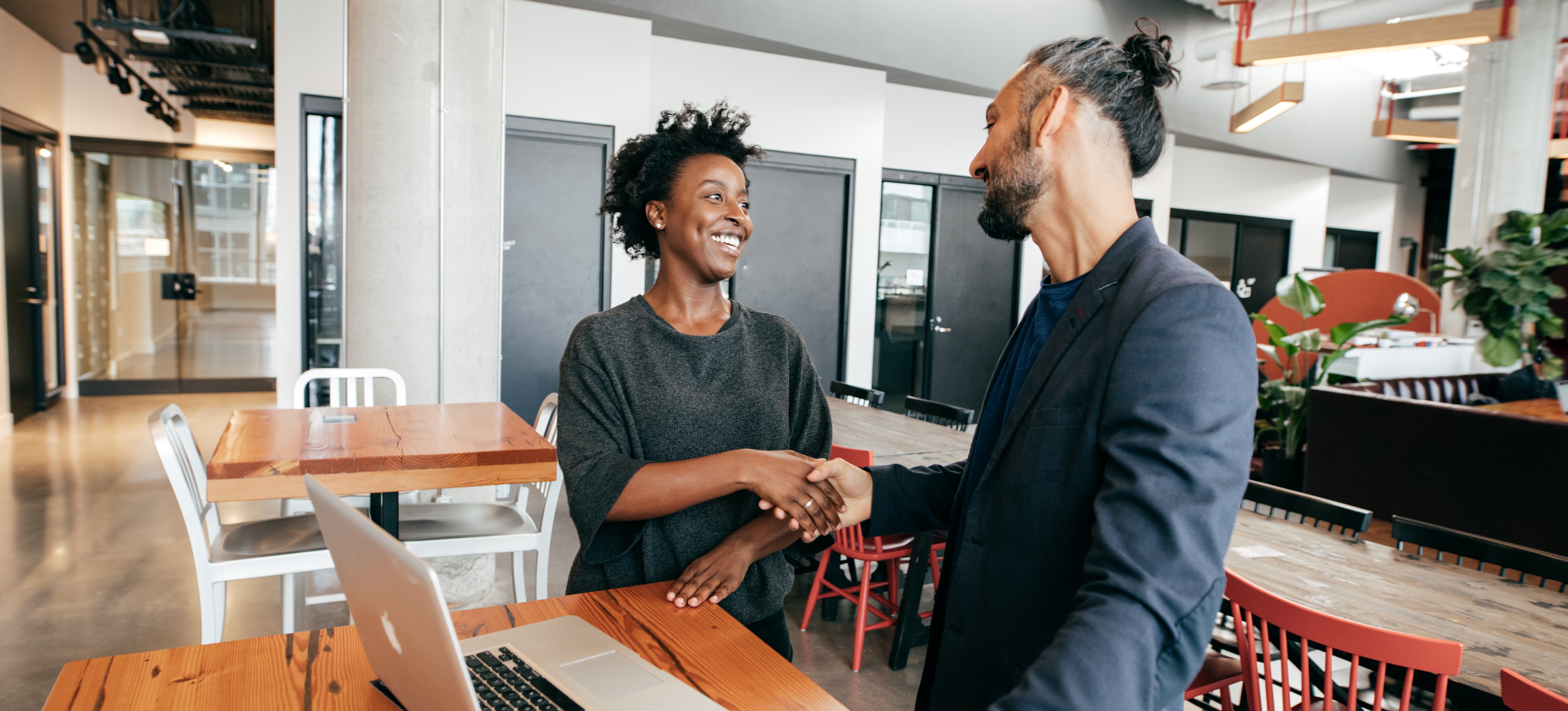 [Featured image] A woman applying for a back-end developer job shakes hands with a hiring manager standing next to a desk in a brightly lit office.
