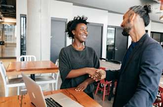 [Featured image] A woman applying for a back-end developer job shakes hands with a hiring manager standing next to a desk in a brightly lit office.