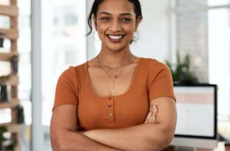 A smiling UX strategist in an orange shirt stands with their arms crossed next to their desk in a design office