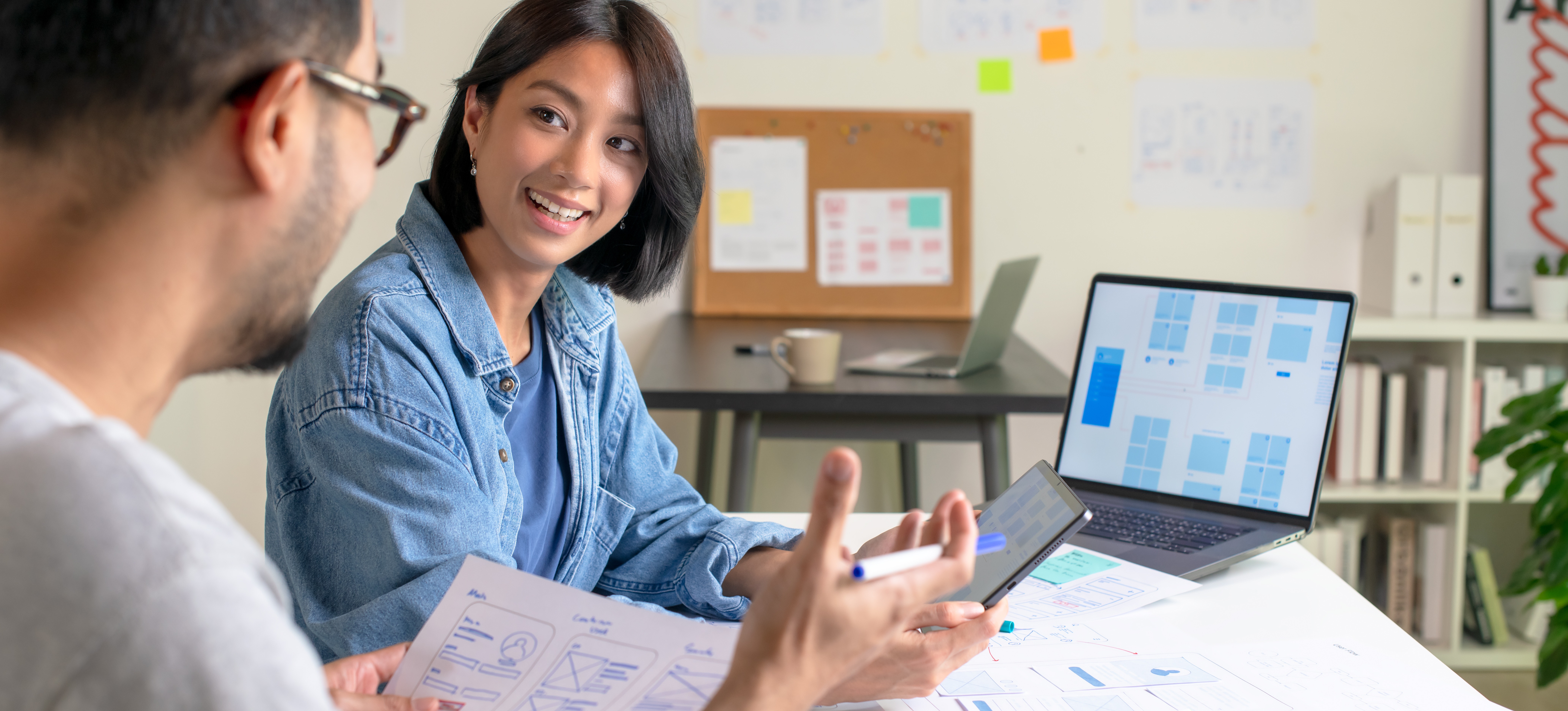 [Featured Image] Two designers are discussing their UX strategy while sitting at a workplace table and looking at designs on paper, a laptop, and a tablet.