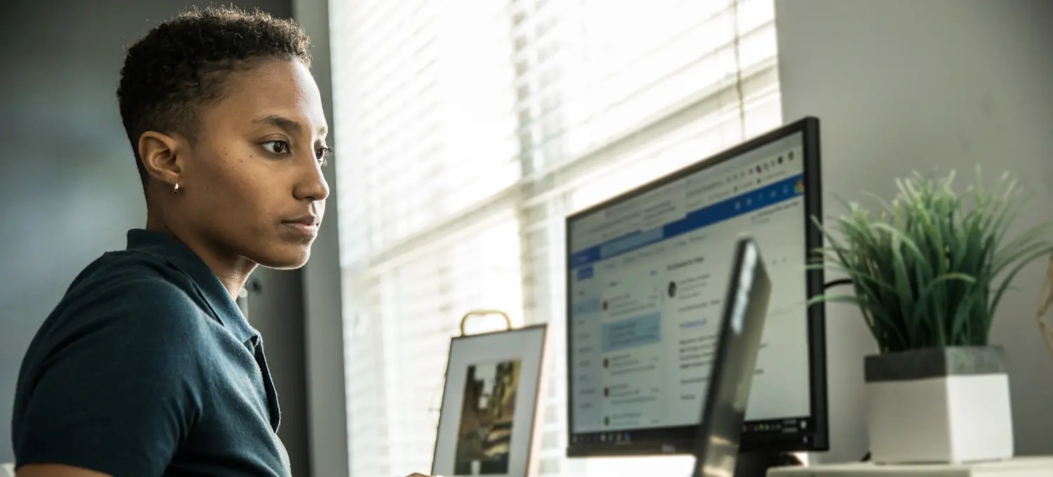 [Featured Image] A data journalist works on their laptop and computer in their office space in front of a window, researching data for their next story.
