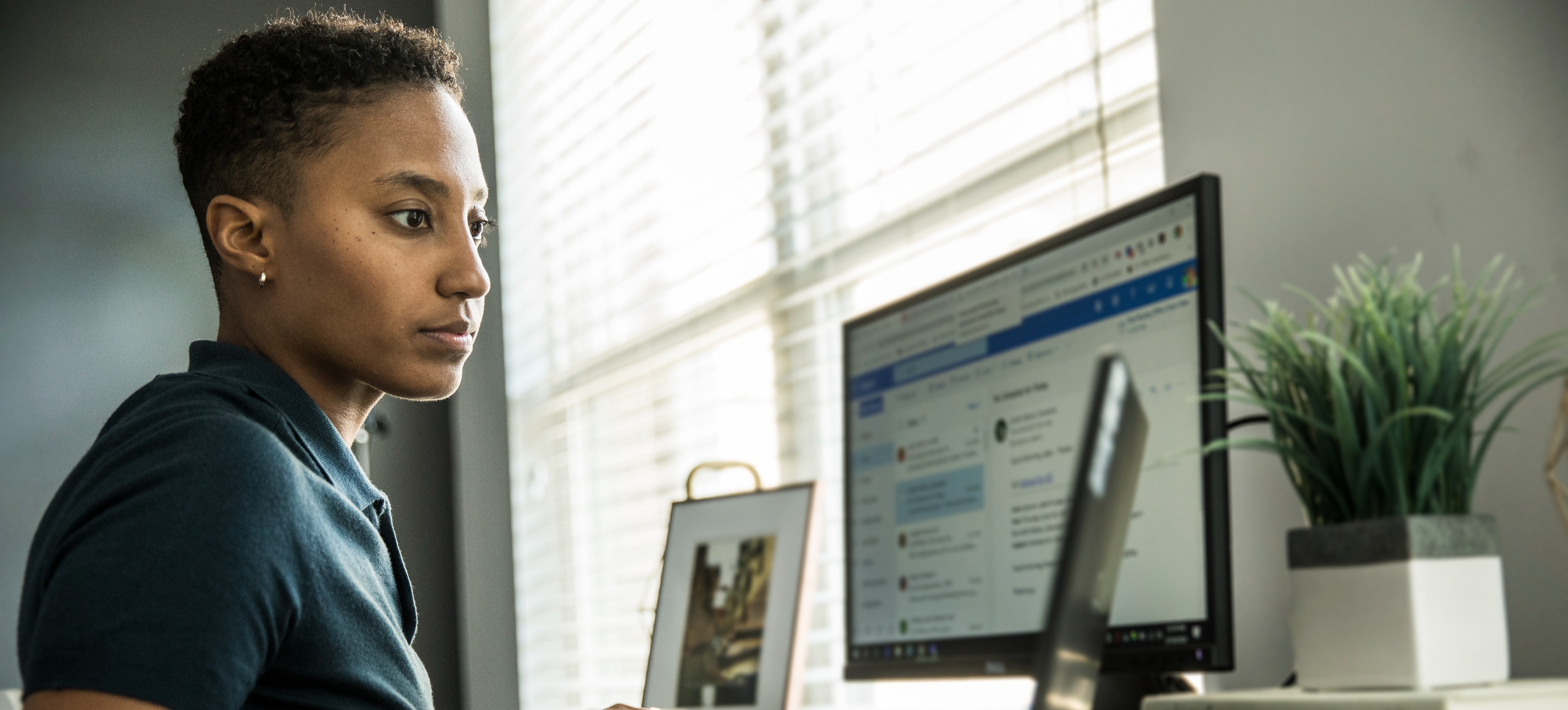 [Featured Image] A data journalist works on her laptop and computer in her office space in front of a window, researching data for her next story.
