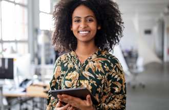 [Featured image] A smiling young woman, wearing a tropical print blouse, stands in a marketing office holding a tablet.