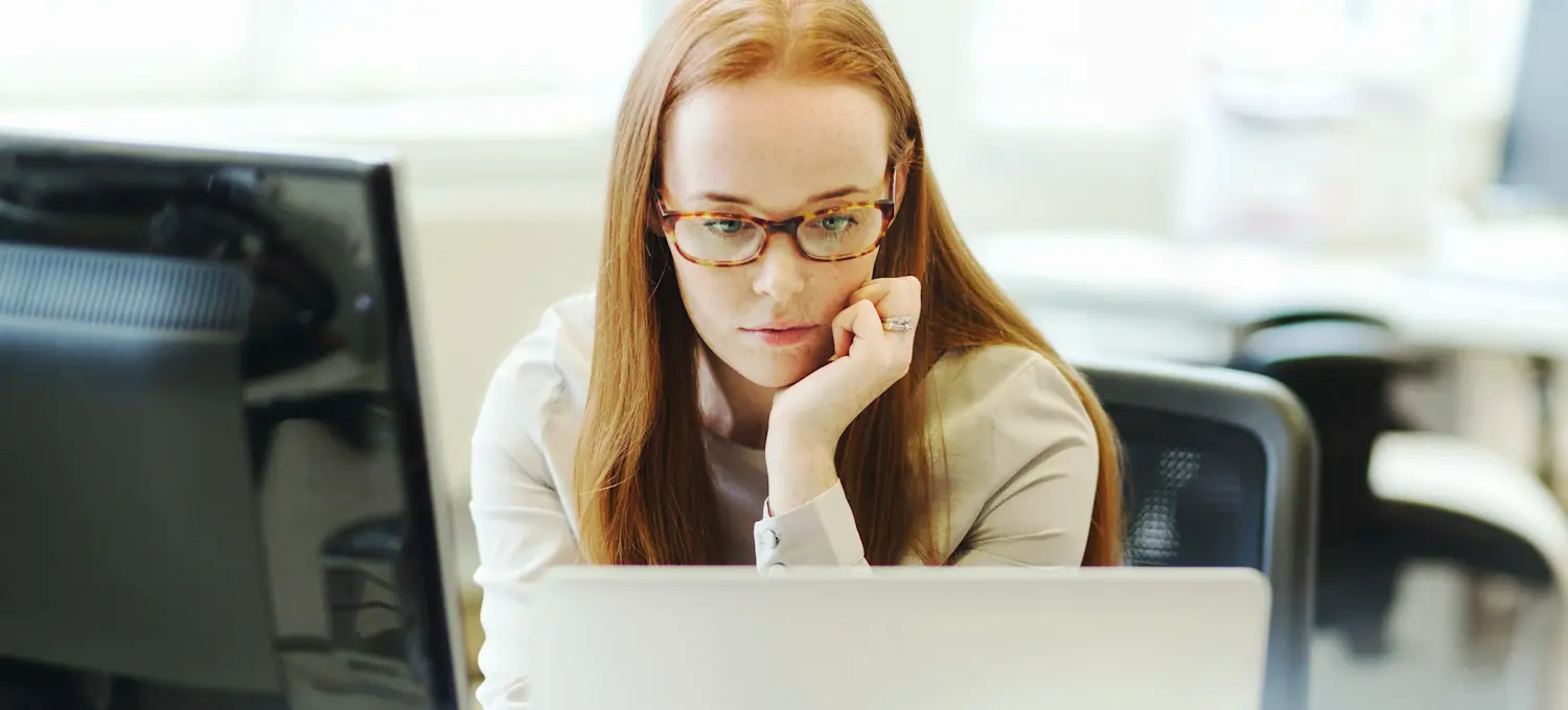 [Featured Image] Young back-end developer with long red hair and glasses looking at two computer screens as they design the server side of a website.
