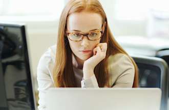 [Featured Image] Young female back-end developer with long red hair and glasses looking at two computer screens as she designs the server side of a website.
