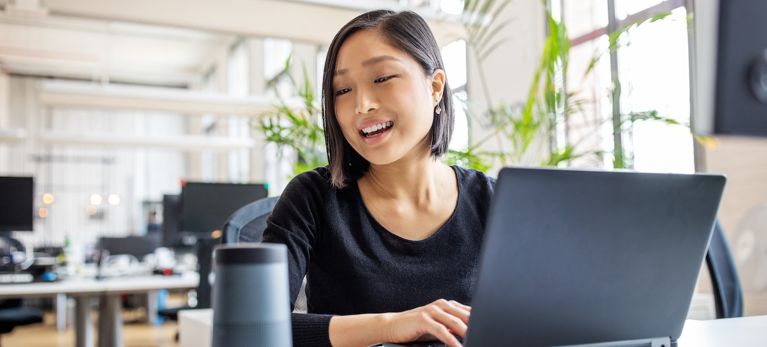 [Featured image] A new employee with an associate degree in business administration sits at their laptop in an open office space, smiling as they prepare for their first day as an executive assistant.