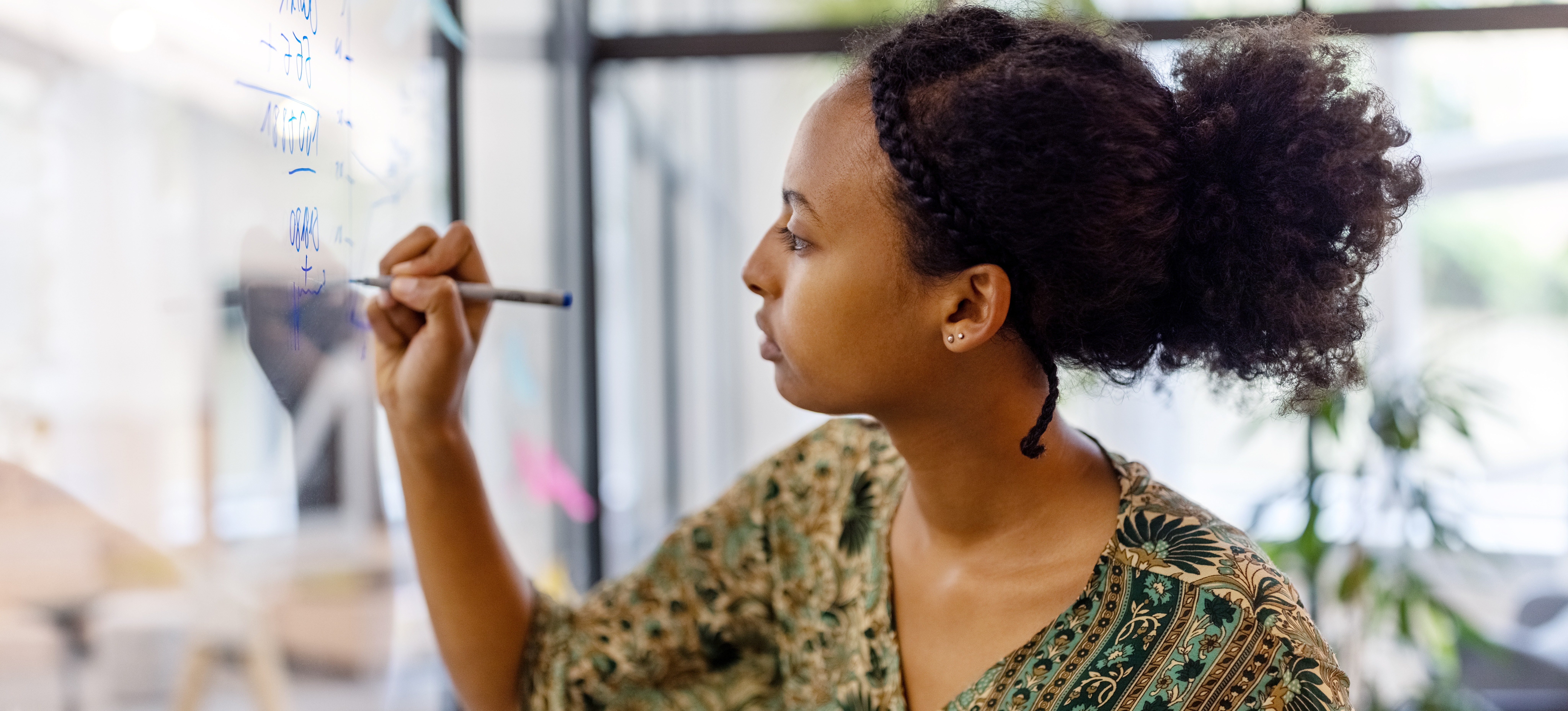 [Featured Image] An economist writes on a glass board and attempts to explain a wicked problem to encourage their colleagues to examine the issue through a new lens. 
