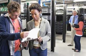 [Featured Image] Coworkers use their cybersecurity skills to assess threats while reviewing information on a clipboard in an office’s technology room. 
