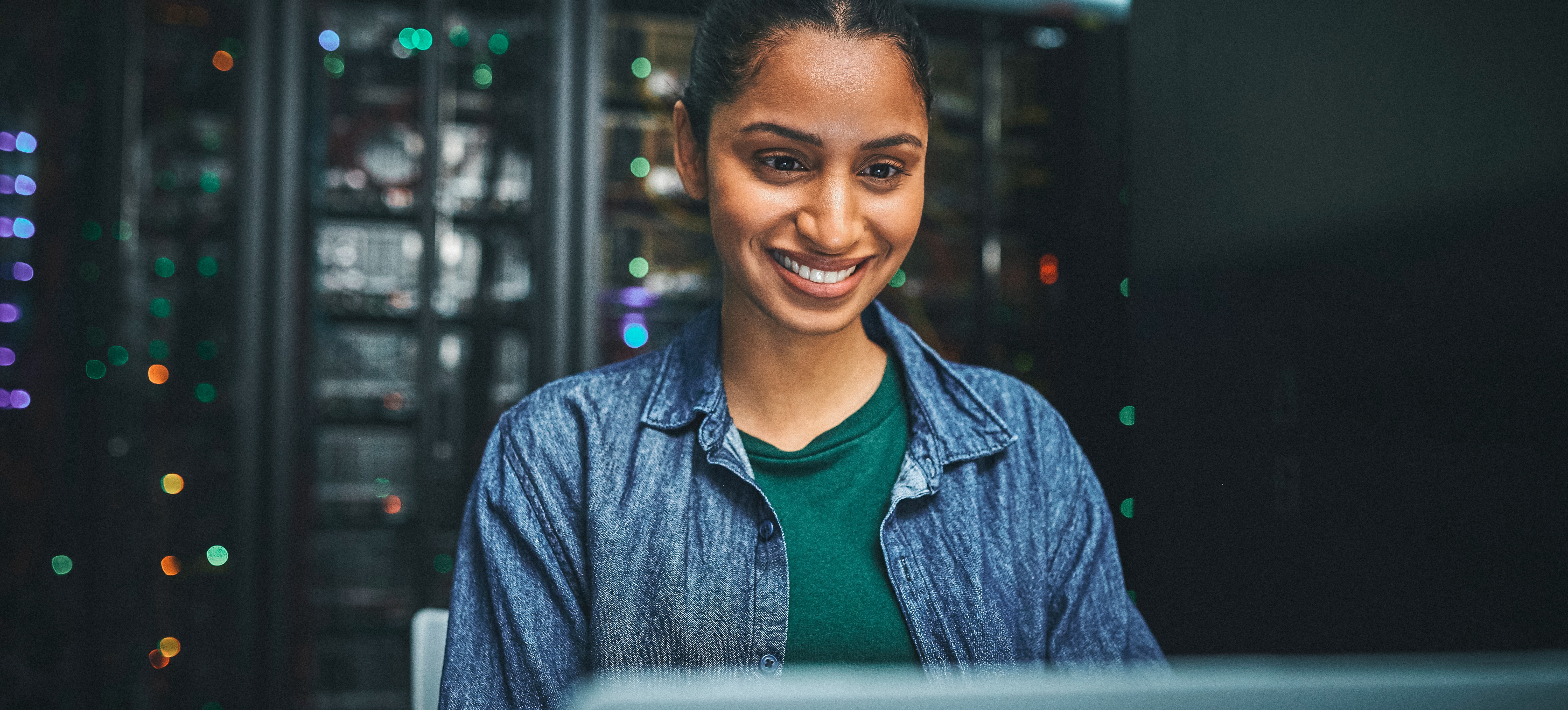 [Featured Image] A smiling cloud computing professional who has earned Google Cloud Certification works at her computer in a server room.
