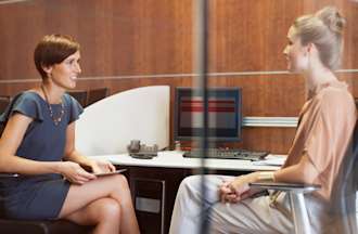 [Featured Image] Two people sit facing each other in a small glass conference room. The person on the left wears a gray dress and the person on the right wears a beige shirt and pants.