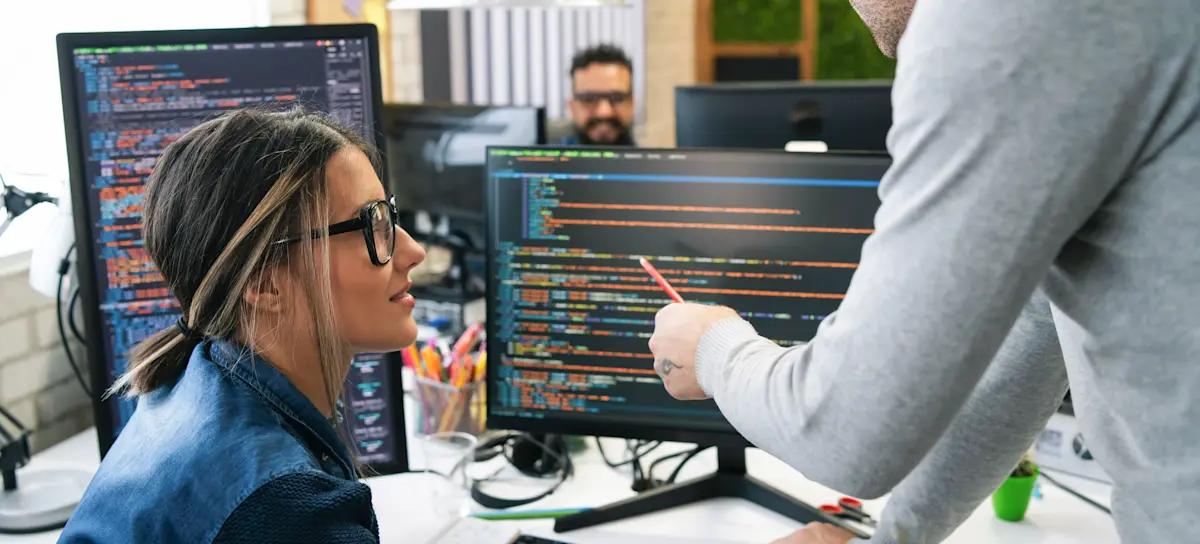 [Featured Image] A developer guiding a colleague through front-end coding on a computer screen in a collaborative workspace.
