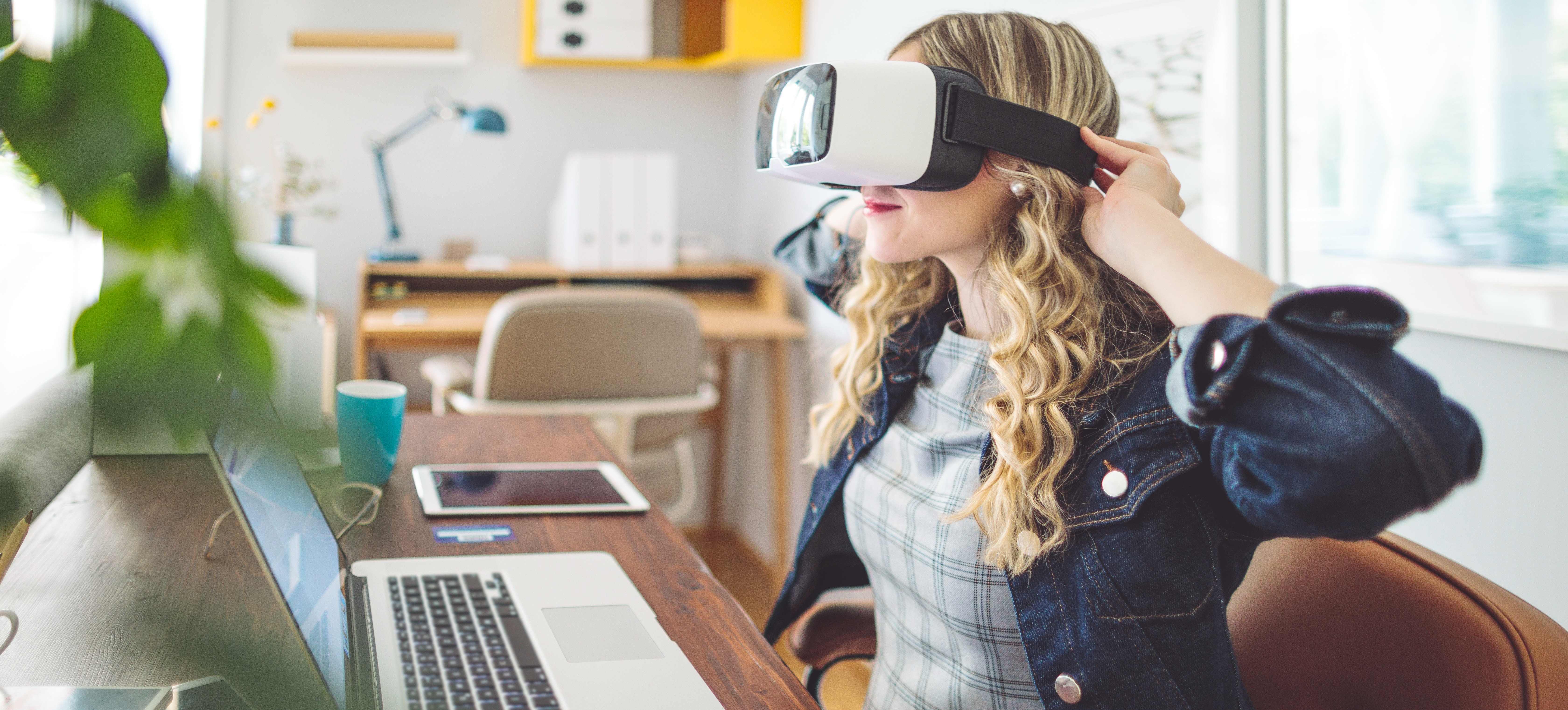 [Featured Image] A video game tester puts on a VR headset as she sits at her desk in front of her laptop.

