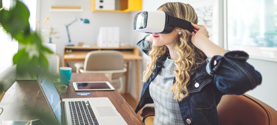 [Featured Image] A video game tester puts on a VR headset as she sits at her desk in front of her laptop.
