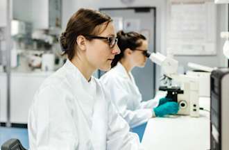 [Featured Image]:  Clinical Research Coordinators wearing lab coats and gloves, sitting in a lab, working on a desktop computer, and using a microscope, to analyze data conducting a clinical trial. 