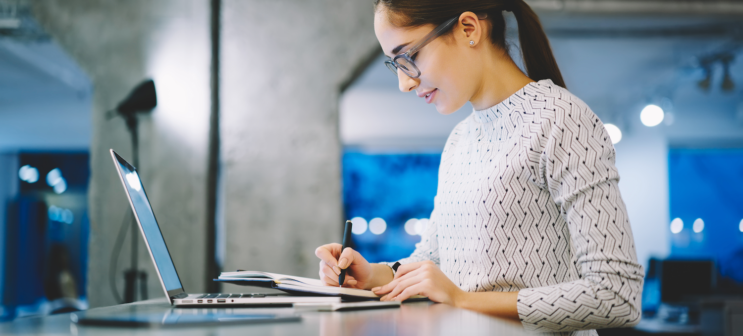 [Image en vedette] Une analyste de données prend des notes sur son ordinateur portable à un bureau debout dans un espace de bureau moderne