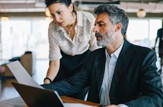 [Featured Image] Two workers are analyzing business plans with a laptop on the desk. 