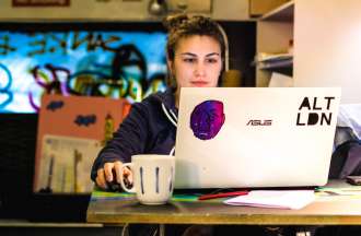 [Featured image] A young woman sits at a table in a cafe writing a resume for her first job on a laptop computer with stickers on it. She has a cup of coffee in front of her on the table.