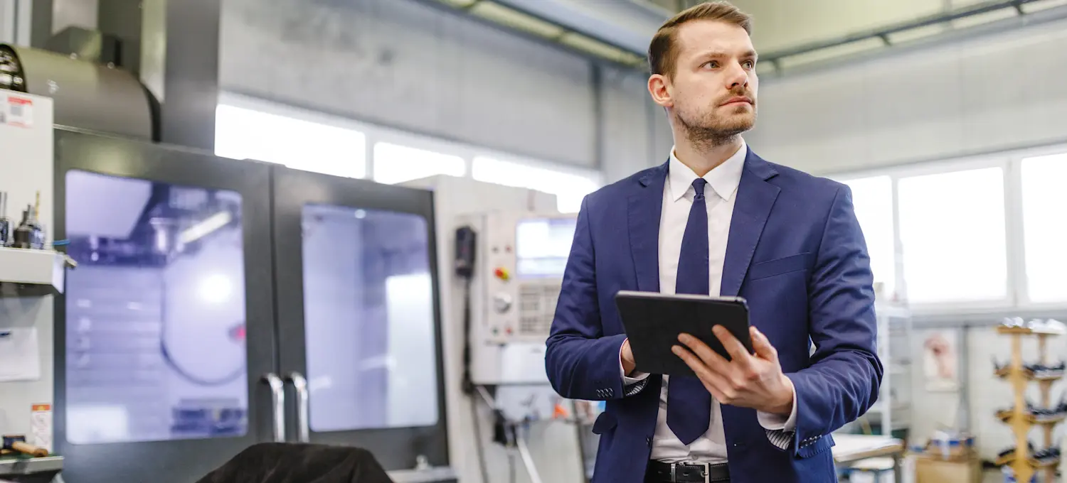[FEATURED IMAGE] A chief product officer in a suit and tie stands in a factory, holding a tablet and observing their surroundings. 
