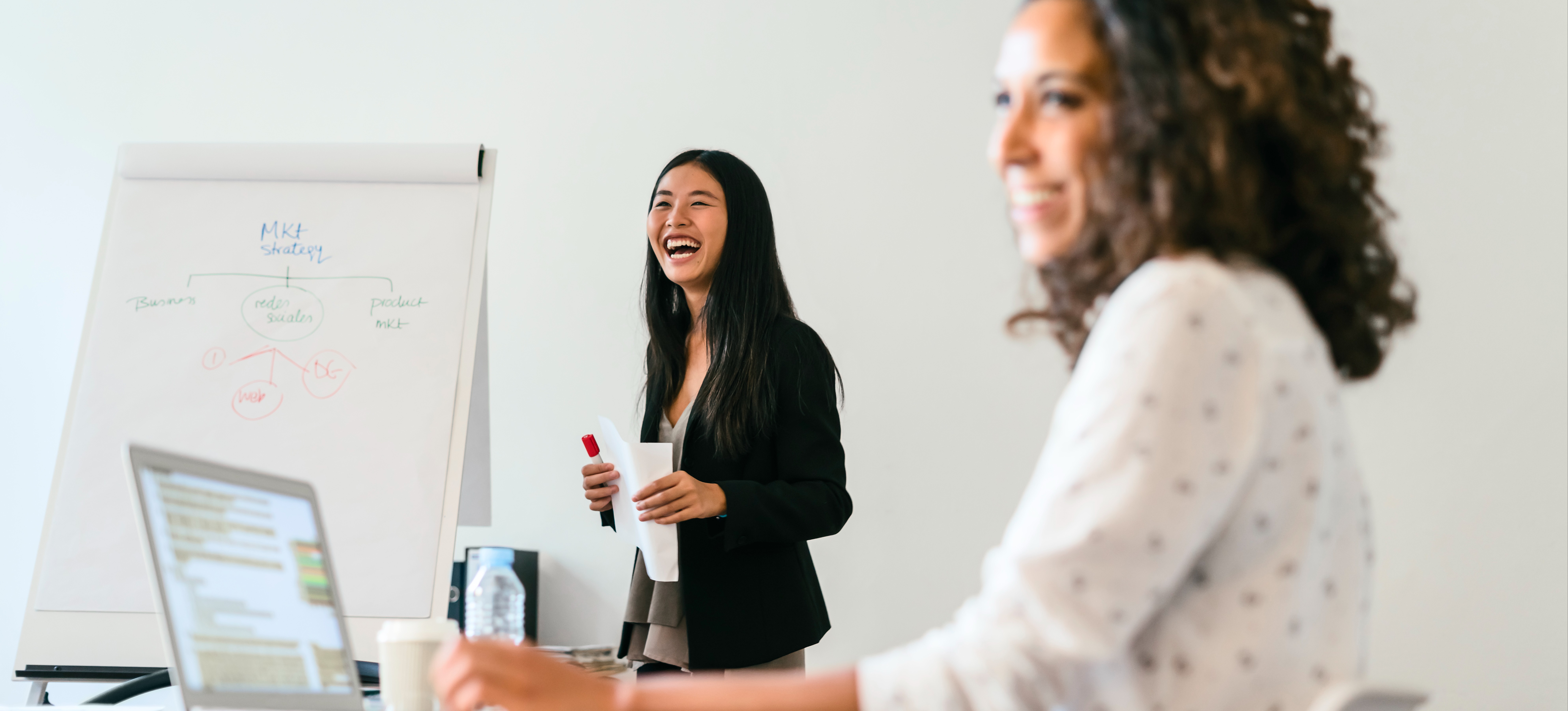 [Featured Image] A laughing marketing specialist presents to other businesspeople in a conference room, using a large flip pad on an easel.
