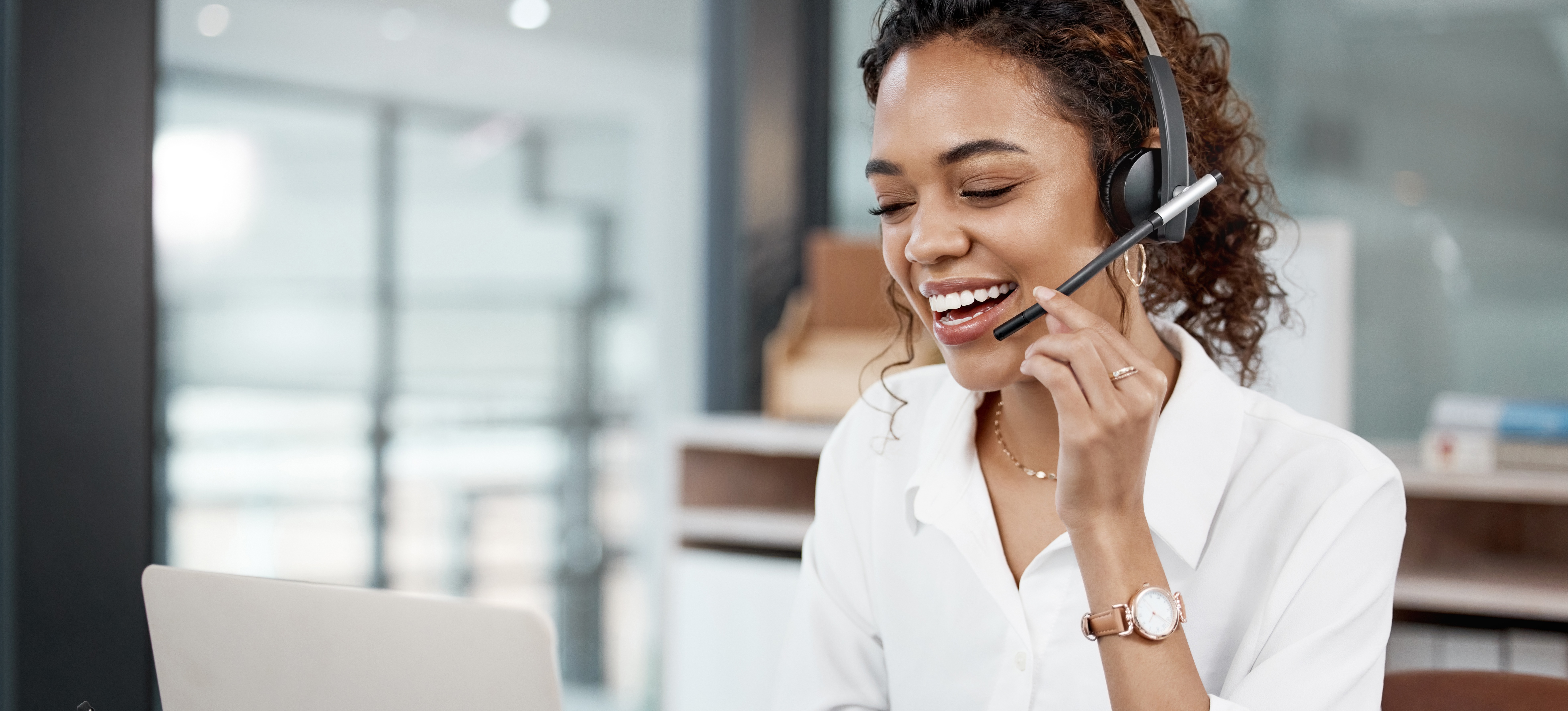 [Featured Image] A medical sales rep sits at a desk and looks at a laptop as she uses a headset to talk to a client.  
