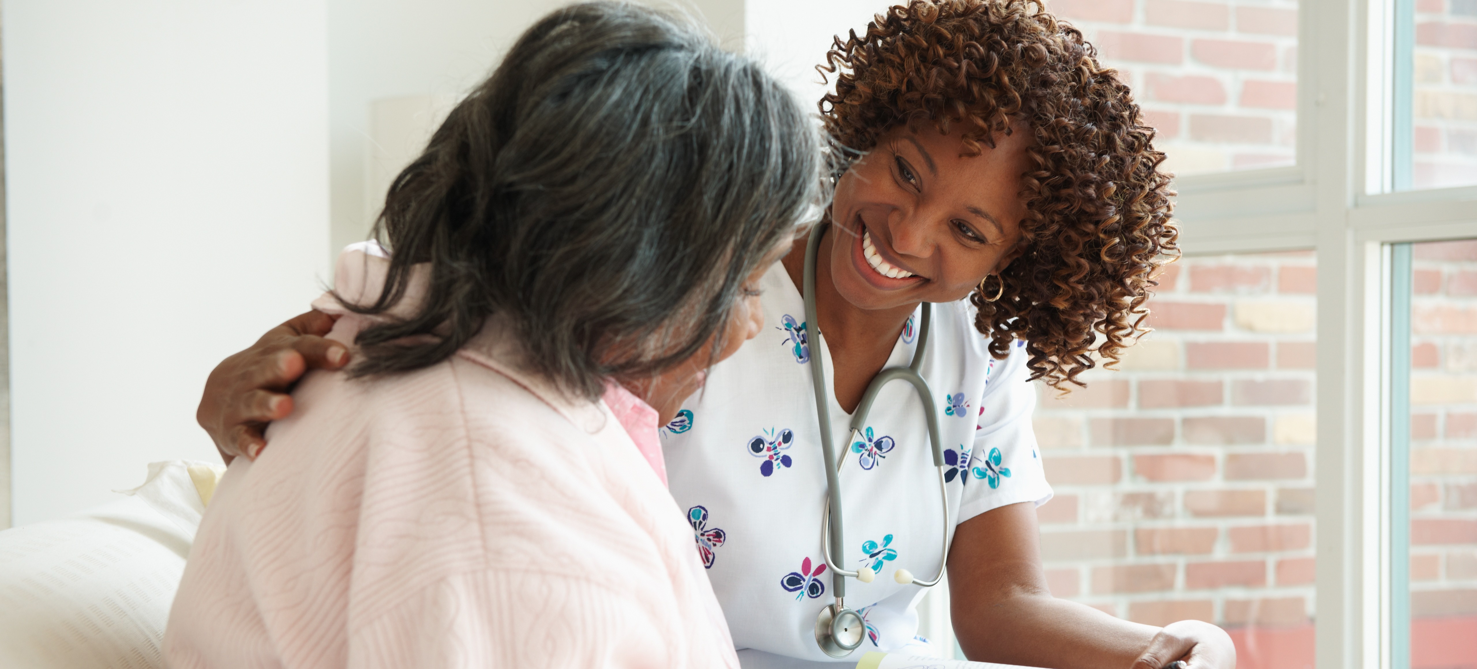 [Feature Image] A hospice nurse smiles at her patient as they sit together on a comfortable sofa near a window.
