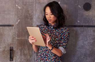 [Featured image] A young woman stands in front of a gray, concrete wall looking at a tablet.