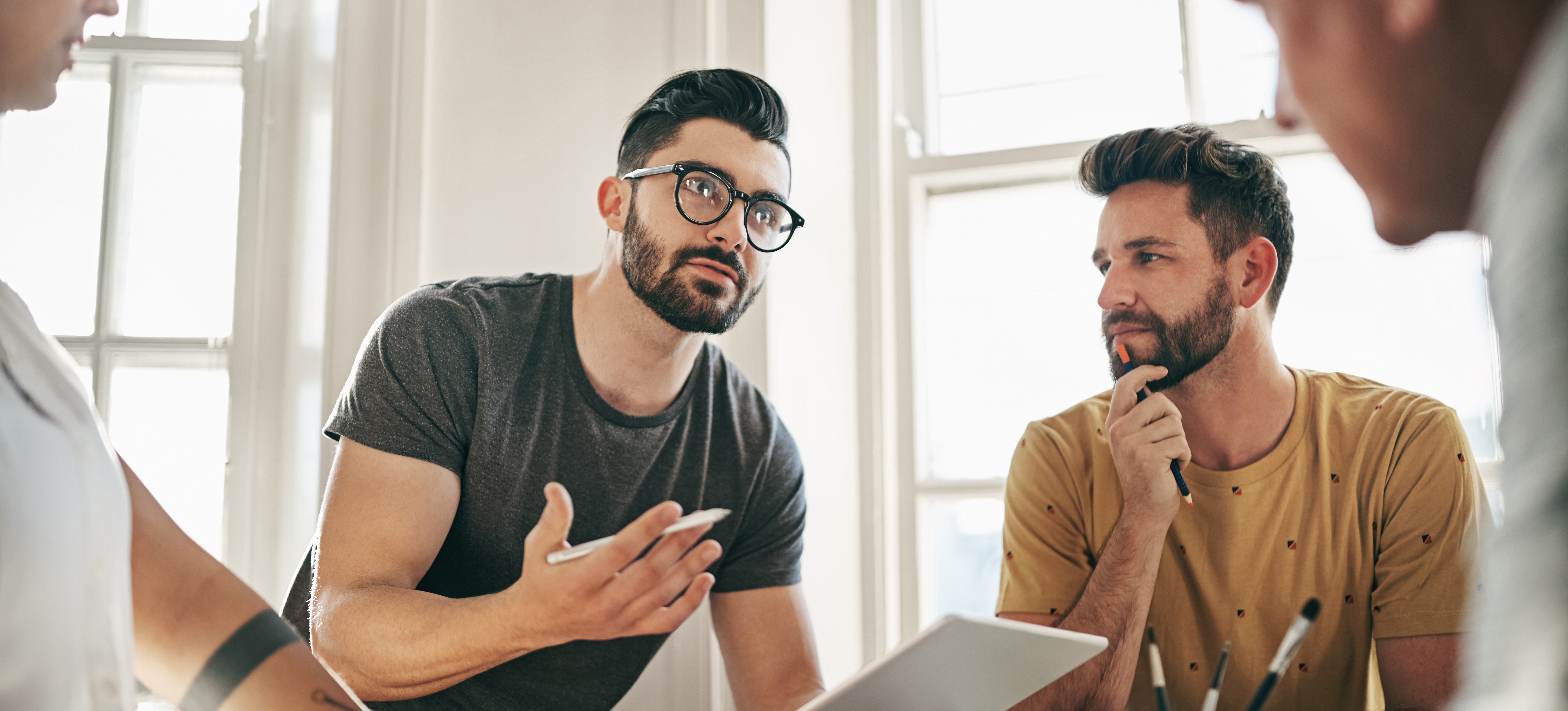 [Featured Image] A group of colleagues discuss a project’s progress, with the project manager using excellent verbal communication skills to convey the strategy moving forward. 
