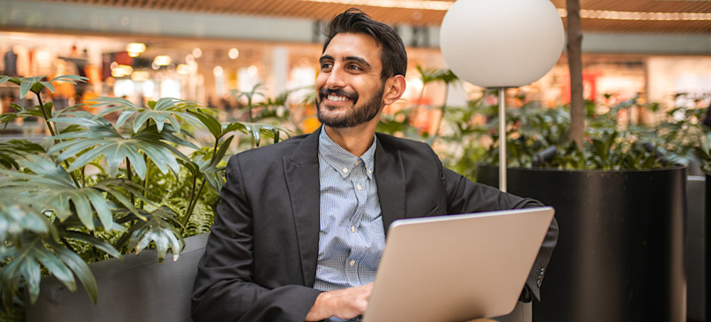 [Featured Image] A smiling professional sits in an outdoor plaza surrounded by greenery as he learns how to write a LinkedIn summary on their laptop. 