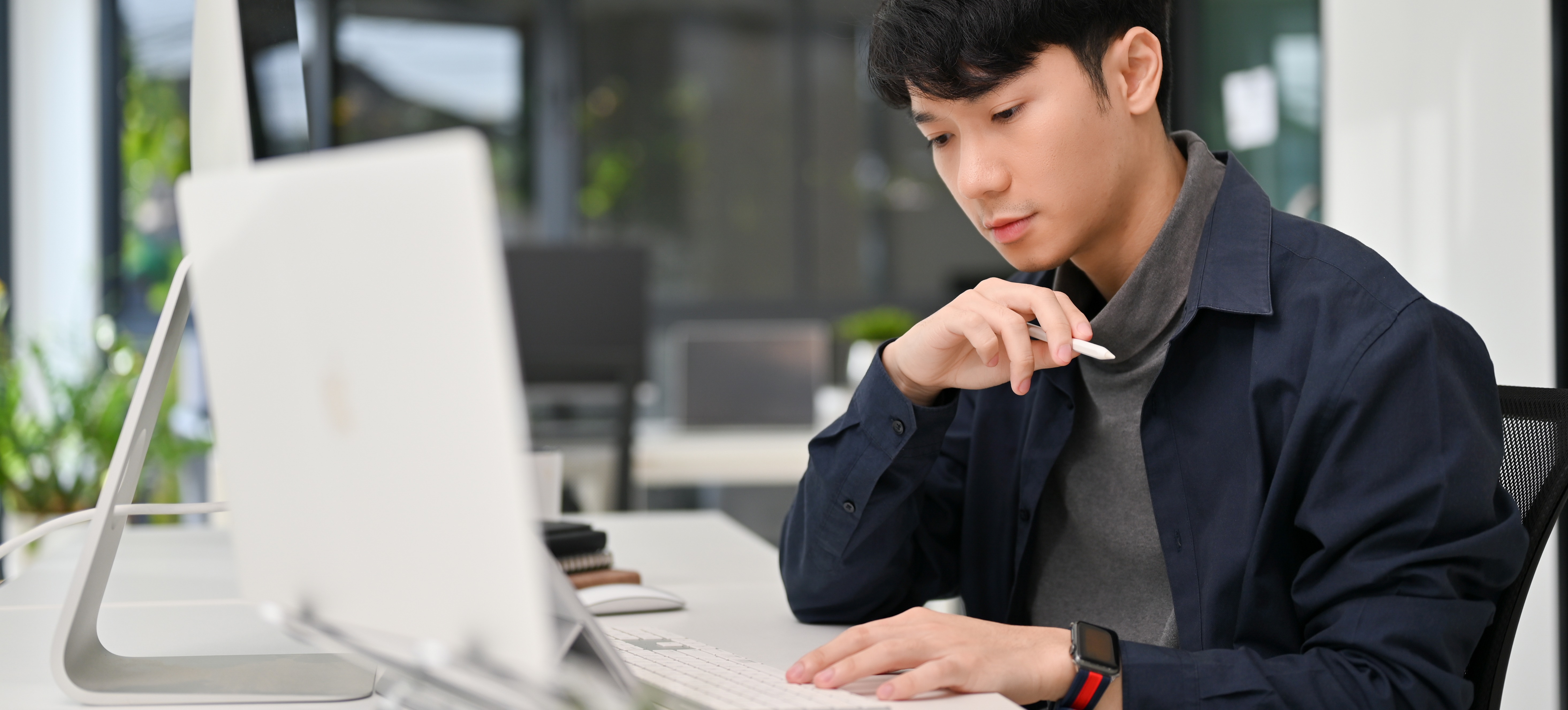 [Featured Image] A young male programmer working on his laptop and a second computer monitor with design patterns in Python in an office setting.
