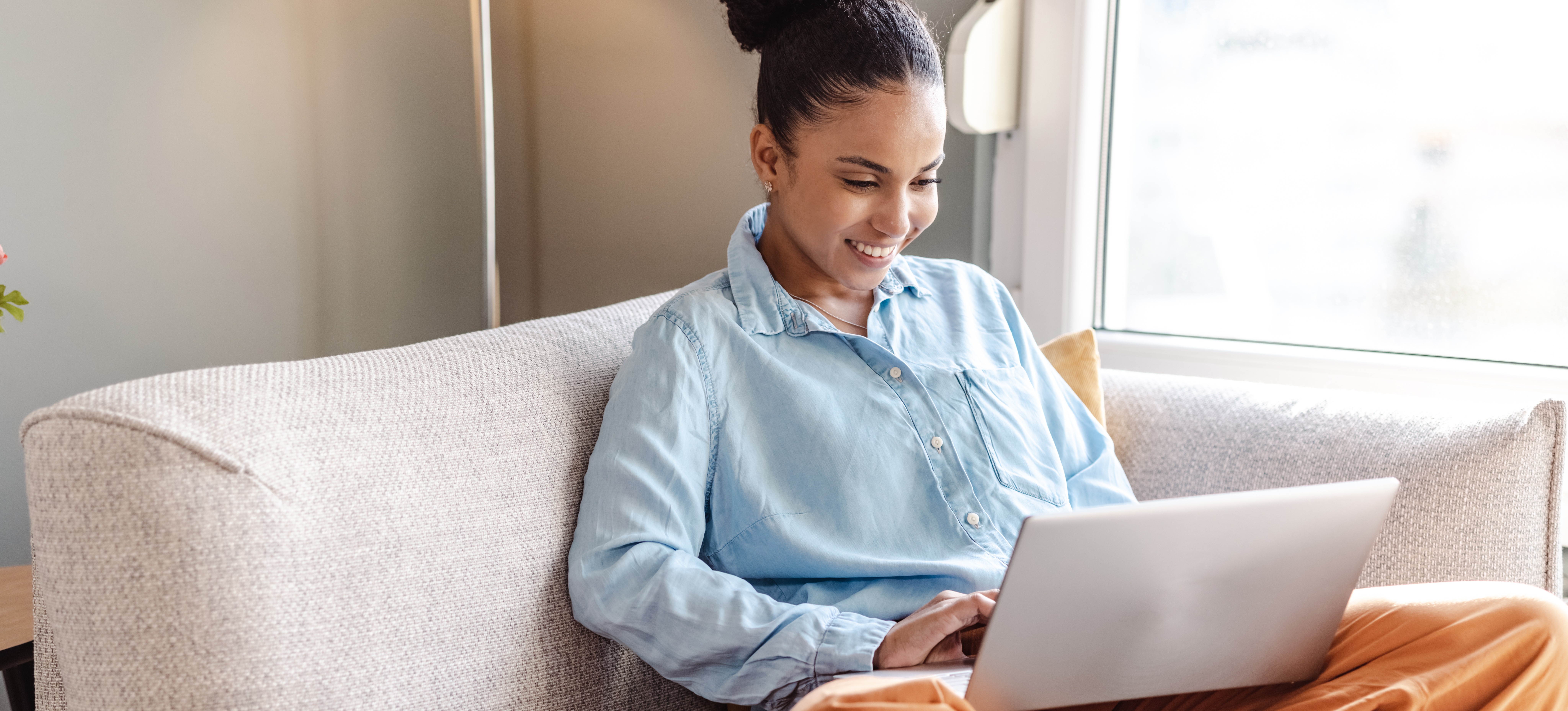 [Featured Image] A woman sits in a chair in her home and researches on her laptop how to apply to a university without GCSEs. 
