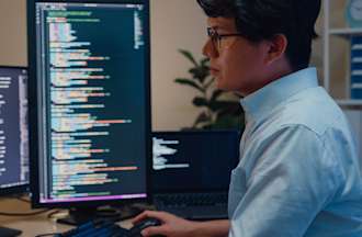 [Featured Image] Young Asian professional male software developer sitting at a desk using three computer monitors to write Python code.