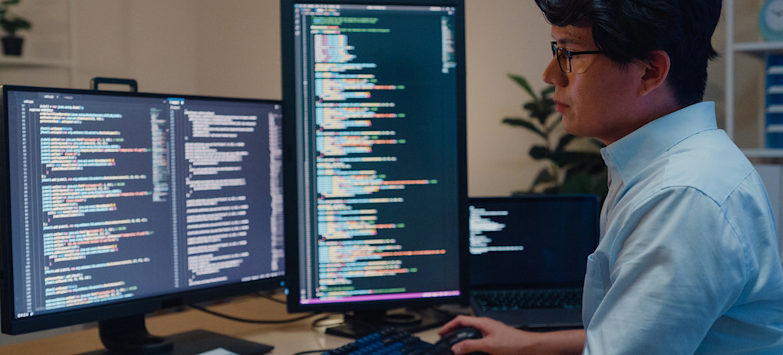 [Featured Image] A business professional software developer sitting at a desk using three computer monitors to write Python code.